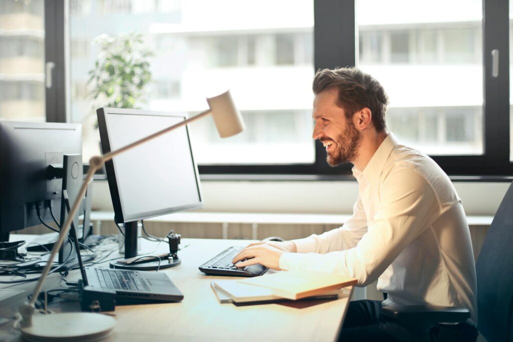 Best Email Hosting for Small Business Success in 2025 8 A man smiling while working at an office desk with a computer and natural daylight streaming in through large windows.