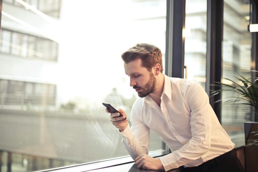 The Essential Guide to Choosing the Right Small Business Consultant 4 A businessman in a white shirt texts on his smartphone by a large glass window inside an office.