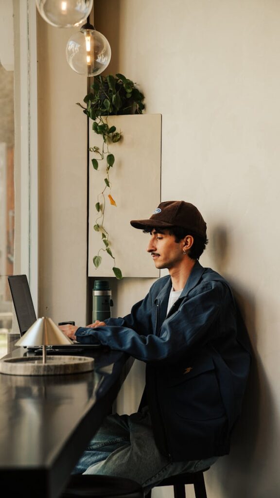 How to Become a Social Media Content Creator in 2025 8 Young man wearing cap and jacket works on laptop in a stylish cafe with warm lighting and green plants.